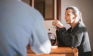 a woman in a grey cardigan drinking coffee at a table in discussion with another person
